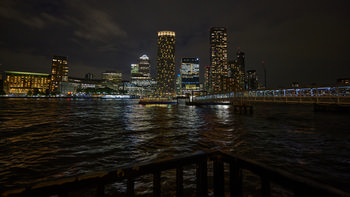 Canary Wharf night pier This urban night photograph shows Canary Wharf in London, United Kingdom, as seen from the pier by the River Thames. The scene was captured in the evening during early autumn, showcasing illuminated skyscrapers such as One Canada Square and other prominent buildings that define the Canary Wharf skyline. Reflections from the office towers and riverside lights shimmer across the water, highlighting the vibrant atmosphere along the Thames. The architecture of the area is clearly visible in the image, with the Canary Wharf pier and modern footbridge stretching out toward the high-rise district, illustrating the dynamic urban environment synonymous with central London.